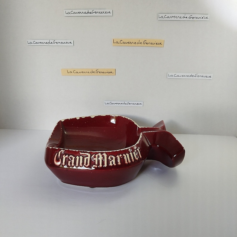May include: A red ceramic ashtray shaped like a bottle with the words "Grand Marnier" in white lettering.