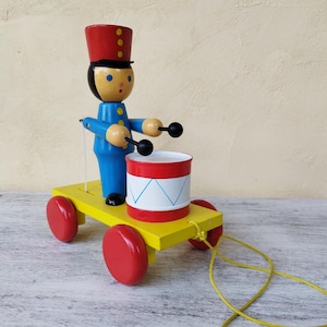 Op de afbeelding: Een vintage houten speelgoed drummer op een gele kar met rode wielen. De drummer draagt een blauw uniform en een rode hoed. De trommel is rood en wit met een blauw en wit ontwerp. Een gele draad is aan de kar bevestigd.