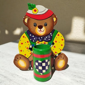May include: A wooden bear-shaped pencil holder with a green and red cylindrical cup. The bear has a brown face, a white and red hat, and a yellow shirt with red hearts. The cup features a checkered heart design.