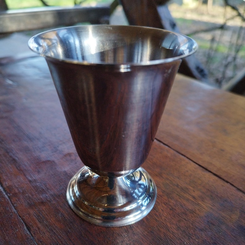 May include: A silver metal goblet with a flared rim and a round base. The goblet is sitting on a wooden surface.