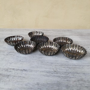 Op de afbeelding: Acht kleine, zilverkleurige metalen bakvormen met geribbelde randen. De vormen zijn in een halve cirkel op een wit houten oppervlak gerangschikt.