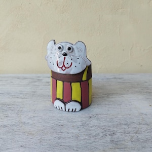 May include: A ceramic cat figurine. The cat's head is white with black eyes and a red mouth. The body is a cylinder with vertical stripes of yellow and red. The cat has white paws.