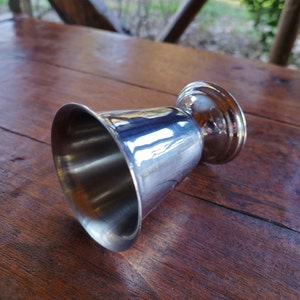 May include: A silver metal jigger with a rounded base and a flared top. The jigger is sitting on a wooden surface.