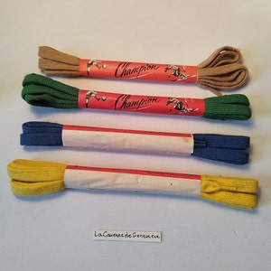 Op de afbeelding: Vier paar veters in verschillende kleuren: bruin, groen, blauw en geel. Elk paar is verpakt in wit papier met rode tekst die "Champion" zegt en een afbeelding van een persoon die voetbal speelt. De tekst "Qualitat Ambra" is ook zichtbaar op het papier.