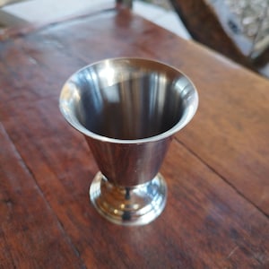 May include: A silver metal cup with a flared rim and a round base. The cup is empty and sitting on a wooden surface.