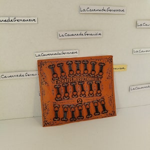 Peut inclure: Une carte avec un fond en cuir brun et des fermoirs en métal noir. La carte indique "AGRAFES PERFECTIONNEES" et "FABRICATION 2 FRANÇAISE".