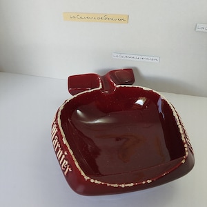 May include: A red ceramic ashtray shaped like a bottle with the word "Marnier" written on the side. The ashtray has a white rim and a white inscription on the side.