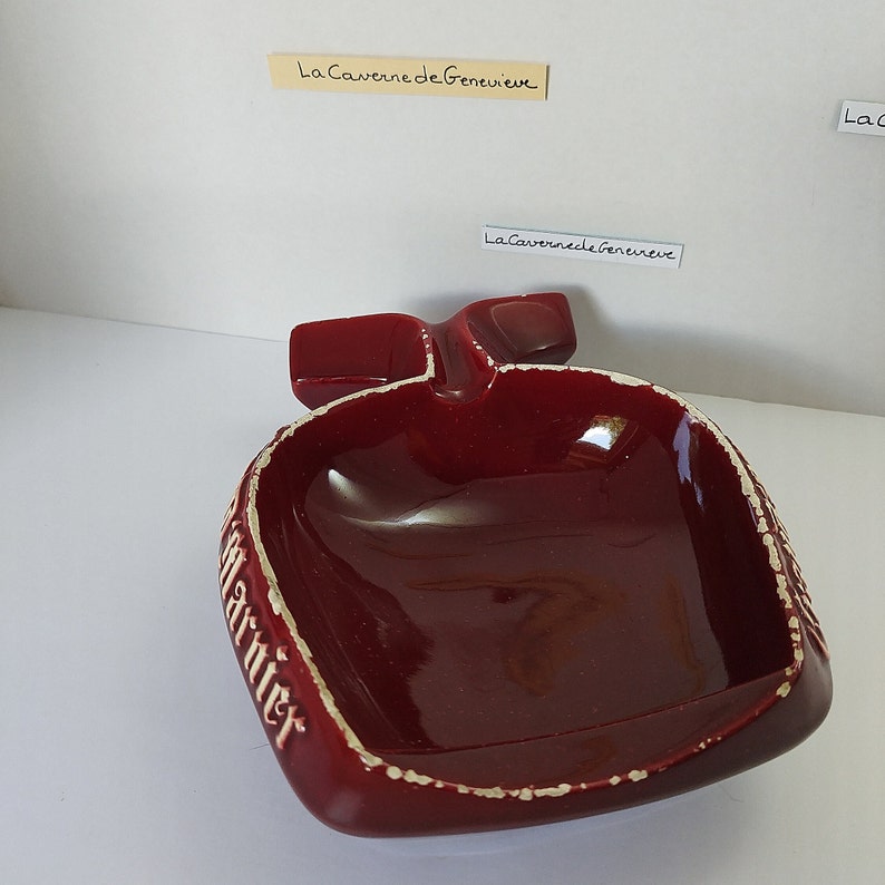 May include: A red ceramic ashtray shaped like a bottle with the word "Marnier" written on the side. The ashtray has a white rim and a white inscription on the side.