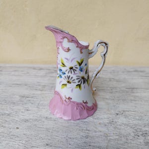 Peut inclure: Un petit pichet en céramique blanche avec une base et un bord roses, orné de bordures dorées et de motifs floraux. Le pichet est doté d'une anse et d'un bec délicats, parfaits pour servir de la crème ou des sauces. Le motif floral comprend des marguerites et d'autres petites fleurs.