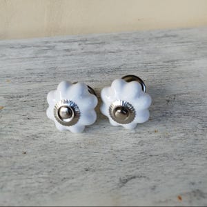 May include: Two white ceramic cabinet knobs with a flower-like design and silver-colored metal accents. These decorative knobs are ideal for adding a touch of elegance to furniture or cabinetry. The knobs are shown on a light-colored, textured surface.
