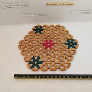 Può includere: Un sottobicchiere rotondo in legno con un motivo geometrico di piccoli pezzi ovali. Il sottobicchiere ha tre accenti a forma di fiore verde e un fiore rosso.