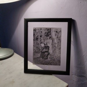 May include: A black framed photo of a woman in a patterned dress sitting in a wooded area.