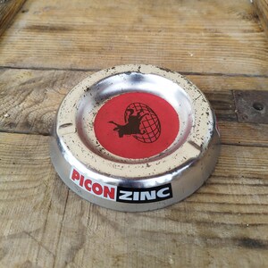 Op de afbeelding: Een zilveren metalen asbak met een rood middenstuk met een zwart stieren- en globeontwerp. De asbak heeft een zwarte sticker op de bodem met de tekst "PICON ZINC".