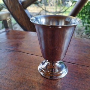 May include: A silver metal goblet with a wide, flared rim and a round base. The goblet is sitting on a wooden surface.