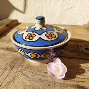 Peut inclure: Un bol en céramique bleu et blanc avec un couvercle, décoré de fleurs orange et jaunes. Le bol est posé sur une surface en bois.
