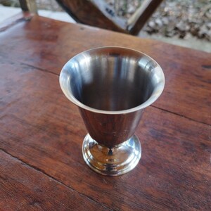 May include: A silver metal cup with a flared rim and a round base. The cup is sitting on a wooden surface.