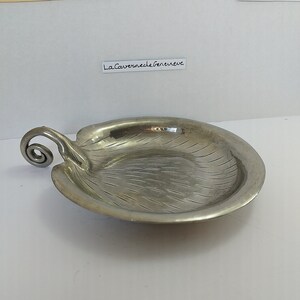 May include: A silver serving dish with a leaf-shaped design and a curled handle. The dish has a textured surface and the text "La Caverne de Geneve" is printed on the side.
