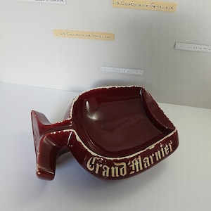 May include: A burgundy ceramic ashtray shaped like a stemmed glass with the words "Grand Marnier" printed in gold on the front.