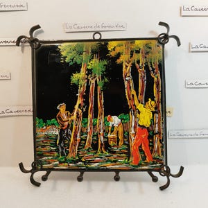 May include: A black metal key holder with a square tile featuring a colorful painting of people tapping maple trees. The tile is framed with a black metal frame and has a decorative design on the bottom.