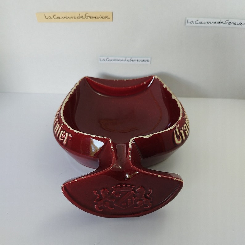 May include: A red ceramic ashtray shaped like a wine glass with a white rim and the words "La Caverne de Geneve" on the side. The bottom of the ashtray has a crest with a star and three lions.