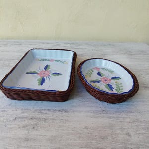 May include: Two serving dishes. One rectangular and one oval, both with white interiors and floral designs in pink, blue, and green. The dishes are surrounded by a brown woven basket-like material.