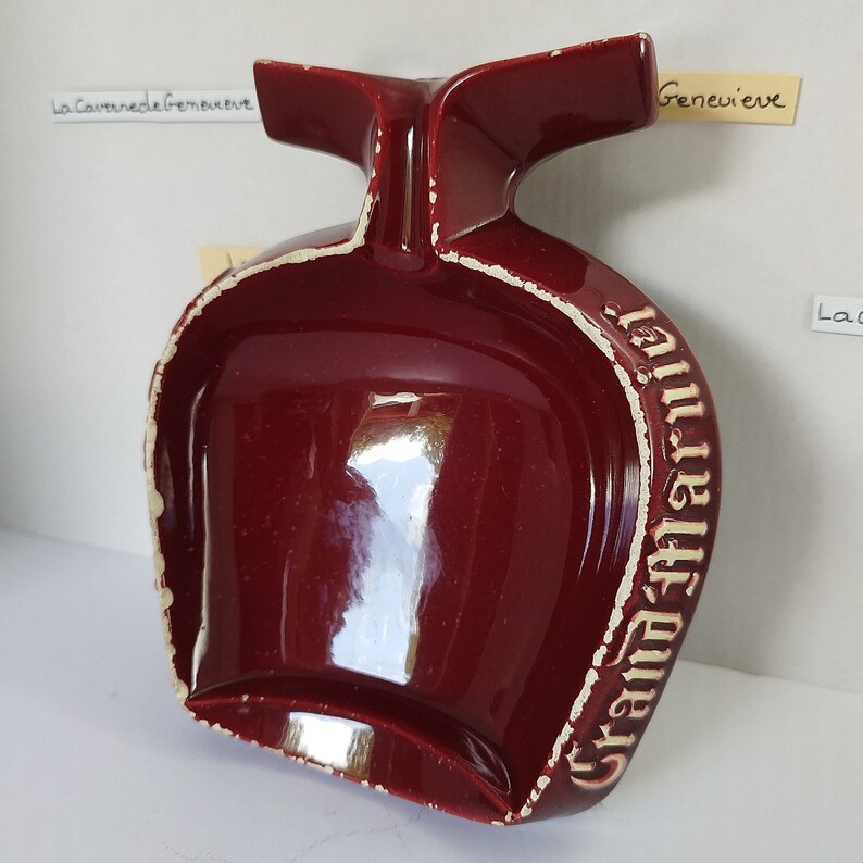 May include: A red ceramic ashtray with a white rim and the words "Grand Marnier" written on the side. The ashtray has a unique shape with a handle on the top.