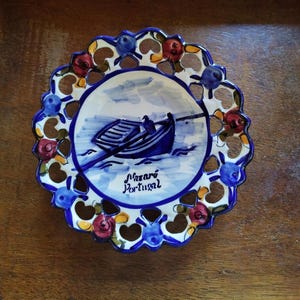 Puede incluir: Un plato de cerámica azul y blanco con un diseño recortado. El plato presenta una pintura de un barco con una persona remando en el agua. El texto "Nazaré Portugal" está escrito en el plato.