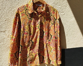 May include: A button-down shirt with a vibrant paisley pattern in shades of yellow, orange, pink, purple, and black. The long-sleeved shirt hangs on a wooden hanger against a textured wall, showcasing its colorful design.