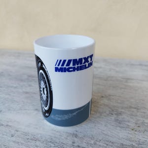 Puede incluir: Taza de cerámica blanca con un gráfico de neumático negro y plateado. La taza tiene el texto azul "MXT MICHELIN" impreso en el lateral. La parte inferior de la taza tiene una banda azul.