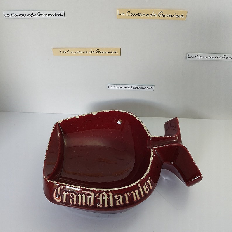 May include: A red ceramic ashtray shaped like a bottle with the words "Grand Marnier" in white lettering.