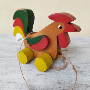 Può includere: Giocattolo di gallo in legno da tirare. Il gallo è dipinto con accenti rossi, verdi, gialli e bianchi. Ha ruote gialle e un cordino. Il giocattolo è in legno e ha un aspetto vintage.