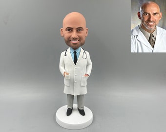 May include: A custom bobblehead figurine of a doctor wearing a white lab coat, a blue tie, and a stethoscope. The figurine is holding a small white cup in one hand.