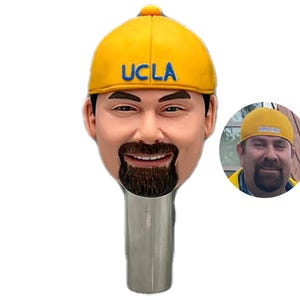 May include: A yellow baseball cap with the text "UCLA" in blue. The cap is on a person's head, and the person's face is visible. The person has a beard and mustache.
