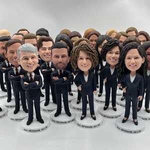 May include: A group of 20 custom bobbleheads, all wearing black suits and white shirts. The bobbleheads are arranged in a semi-circle on a white surface. Each bobblehead has a unique face and hairstyle. The bobbleheads are all facing forward. The text "Sector - GSK Partnership - July 2, 2021" is printed on the base of each bobblehead.