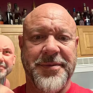 Op de afbeelding: Een close-up van een man met een geschoren hoofd en een grijs baard. Hij draagt een rood shirt. Een kleinere versie van zijn gezicht is zichtbaar op de voorgrond, en lijkt een 3D-geprint model te zijn.