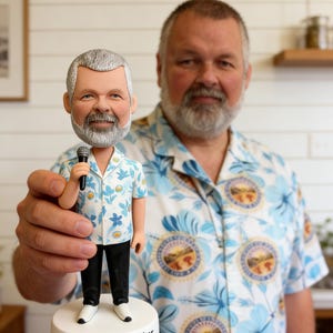 May include: A bobblehead figurine of a man holding a microphone, wearing a floral shirt, black pants, and white shoes. The base reads "2016 Emcee of the Year." The figurine is held in front of a man wearing a similar shirt.
