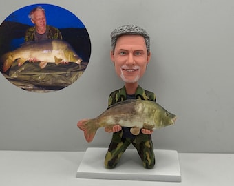 May include: A custom bobblehead figurine of a man holding a large fish. The figurine is wearing camouflage clothing and has a realistic paint job. The man is smiling and has gray hair. The fish is brown and white with a large mouth.
