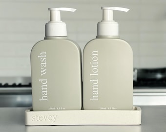 May include: Two gray pump bottles with white labels on a white tray. The bottle on the left says "hand wash" and the bottle on the right says "hand lotion". Both bottles are labeled with "250ml / 8.5 fl oz". The tray has the word "stevey" printed on it.