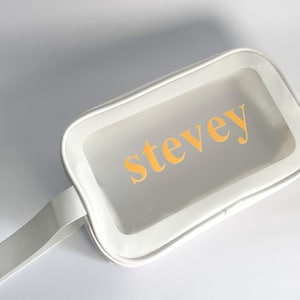 May include: White zippered travel bag with a clear window and a white handle. The bag has the name "stevey" printed in yellow letters.
