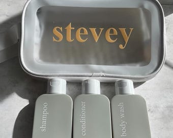May include: Three travel-sized bottles of toiletries in a white zippered pouch. The bottles are labeled "shampoo", "conditioner", and "body wash". The pouch has the word "stevey" printed on it in gold letters.