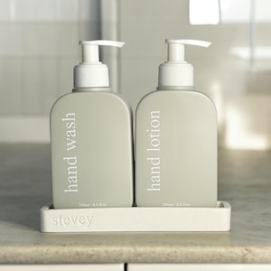 May include: Two grey plastic pump bottles with white labels. The bottle on the left says "hand wash" and the bottle on the right says "hand lotion". Both bottles are labelled with "250ml / 8.5 fl oz". The bottles are sitting on a white rectangular tray with the word "stevey" printed on the front.