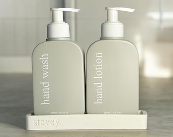 May include: Two gray plastic pump bottles with white labels. The bottle on the left says "hand wash" and the bottle on the right says "hand lotion". Both bottles are labeled with "250ml / 8.5 fl oz". The bottles are sitting on a white rectangular tray with the word "stevey" printed on the front.