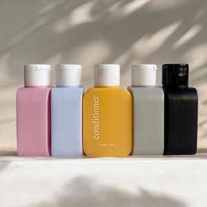 May include: Five small plastic bottles with white caps. The bottles are filled with different coloured liquids. One bottle is labelled "conditioner" and has a yellow liquid. The other bottles have pink, blue, grey, and black liquids.