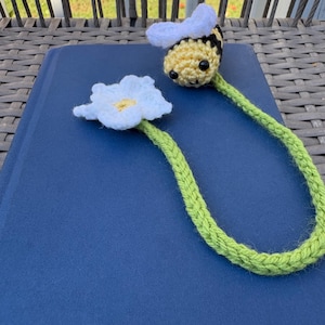 Puede incluir: Un marcapáginas de ganchillo con una abeja de rayas amarillas y negras con alas blancas en un extremo y una flor blanca con centro amarillo en el otro, unidos por un largo tallo de hilo verde trenzado sobre una cubierta de libro azul oscuro.