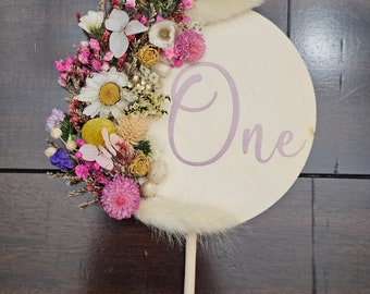 Könnte beinhalten: Ein rundes, weißes Holzschild mit dem Wort "One" in lila Schrift. Das Schild ist mit getrockneten Blumen in den Farben Rosa, Lila, Weiß und Gelb verziert. Das Schild ist an einem Holzstab befestigt.