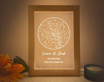 May include: A wooden framed light-up constellation print with a white circle depicting the night sky with stars. The text "Lauren & Jacob" is written below the circle, followed by "Our First Kiss 08.23.2022 | Seattle, WA".