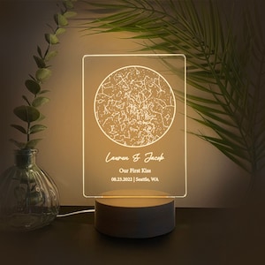 May include: A personalized night light with a wooden base and a clear acrylic panel. The panel displays a constellation map with the names "Lauren & Jacob", the text "Our First Kiss", and the date 08.23.2022, Seattle, WA.