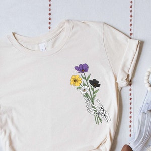 May include: A beige t-shirt with a graphic of a skeleton hand holding a bouquet of purple, yellow, and black flowers.