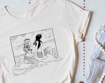 May include: A white t-shirt with a black and white illustration of two mermaids sitting on a rock with their tails in the water. The mermaids are facing away from each other and have long flowing hair.