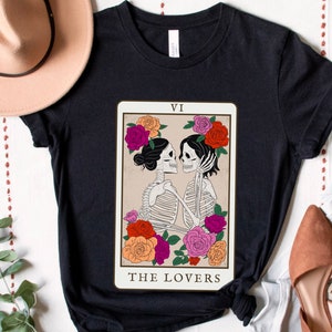 Peut inclure: T-shirt noir avec un motif de carte de tarot repr&eacute;sentant deux squelettes qui s'embrassent. La carte est intitul&eacute;e "The Lovers" et porte le chiffre romain VI en haut.